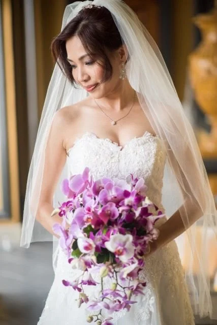 Bride in a wedding dress holding a bouquet of pink and purple orchids, wearing a veil and pearl jewelry, standing indoors.  featuring professional wedding hair and makeup by international stylist Cecilia