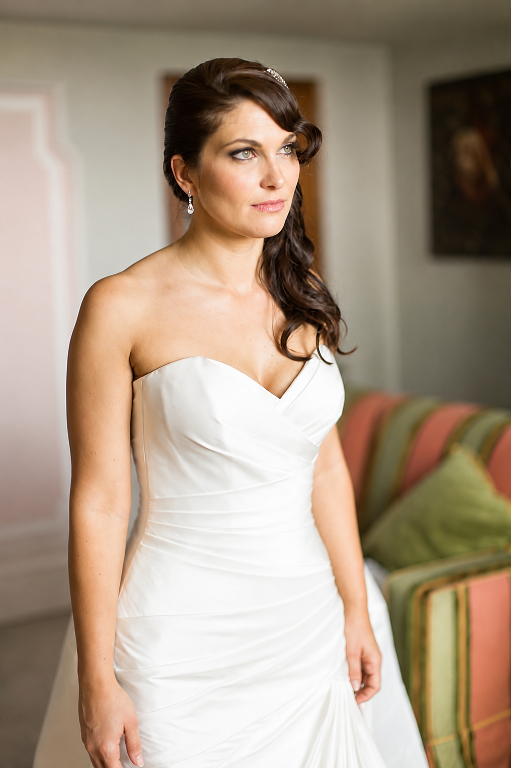 A bride with long dark hair styled in loose curls, and classic makeup by the one and only Cecilia Acquaroli.. is wearing a strapless white wedding gown with a sweetheart neckline, standing in a room with blurred background of sofas and other people.