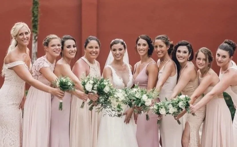 A bride and her nine bridesmaids holding bouquets, smiling in front of a red wall. All styled by Cecilia Acquaroli in Tuscany