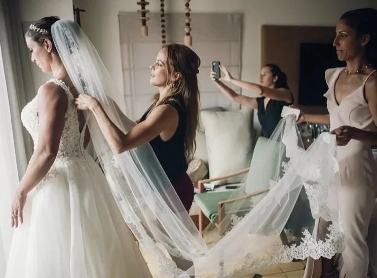 A bride preparing for her wedding, surrounded by women helping with her veil and dress, in a well-lit room.