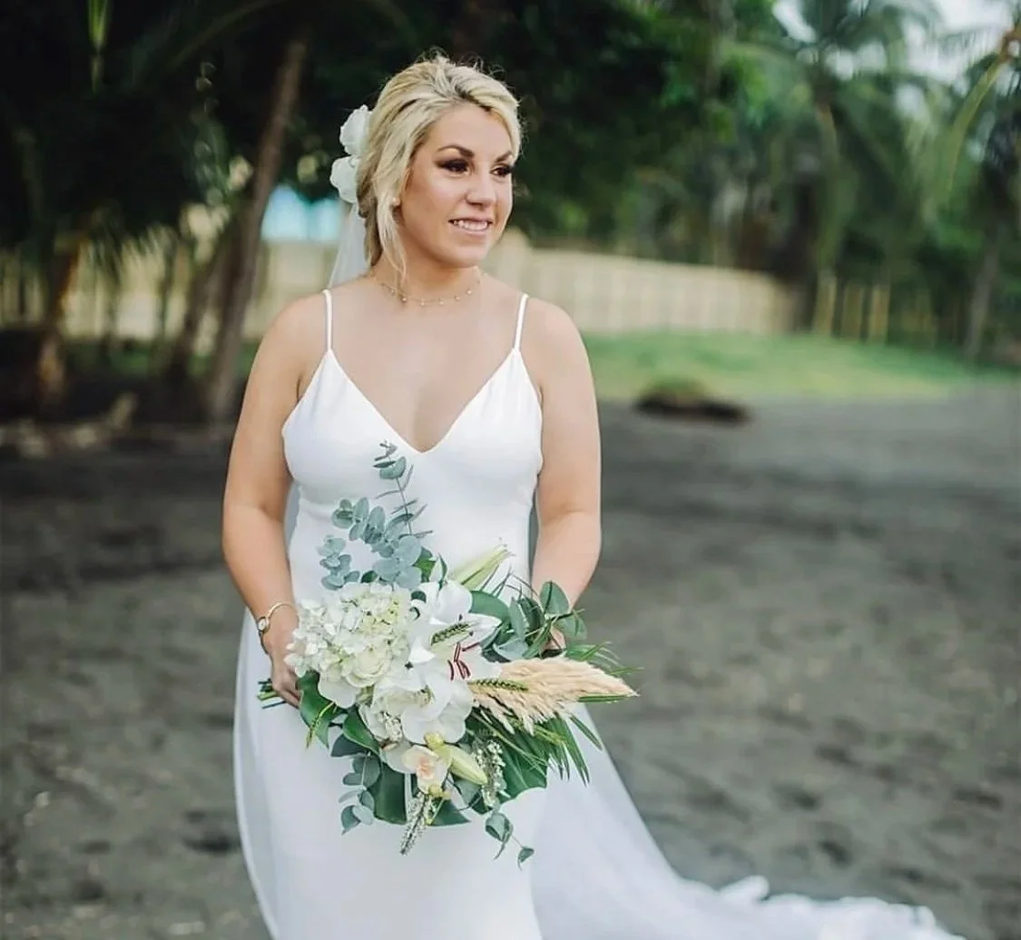 A beach wedding, a styled bride in a white wedding dress holding a bouquet of white flowers and eucalyptus, standing outdoors with trees in the background. Hair and makeup in Italy by Cecilia Acquaroil