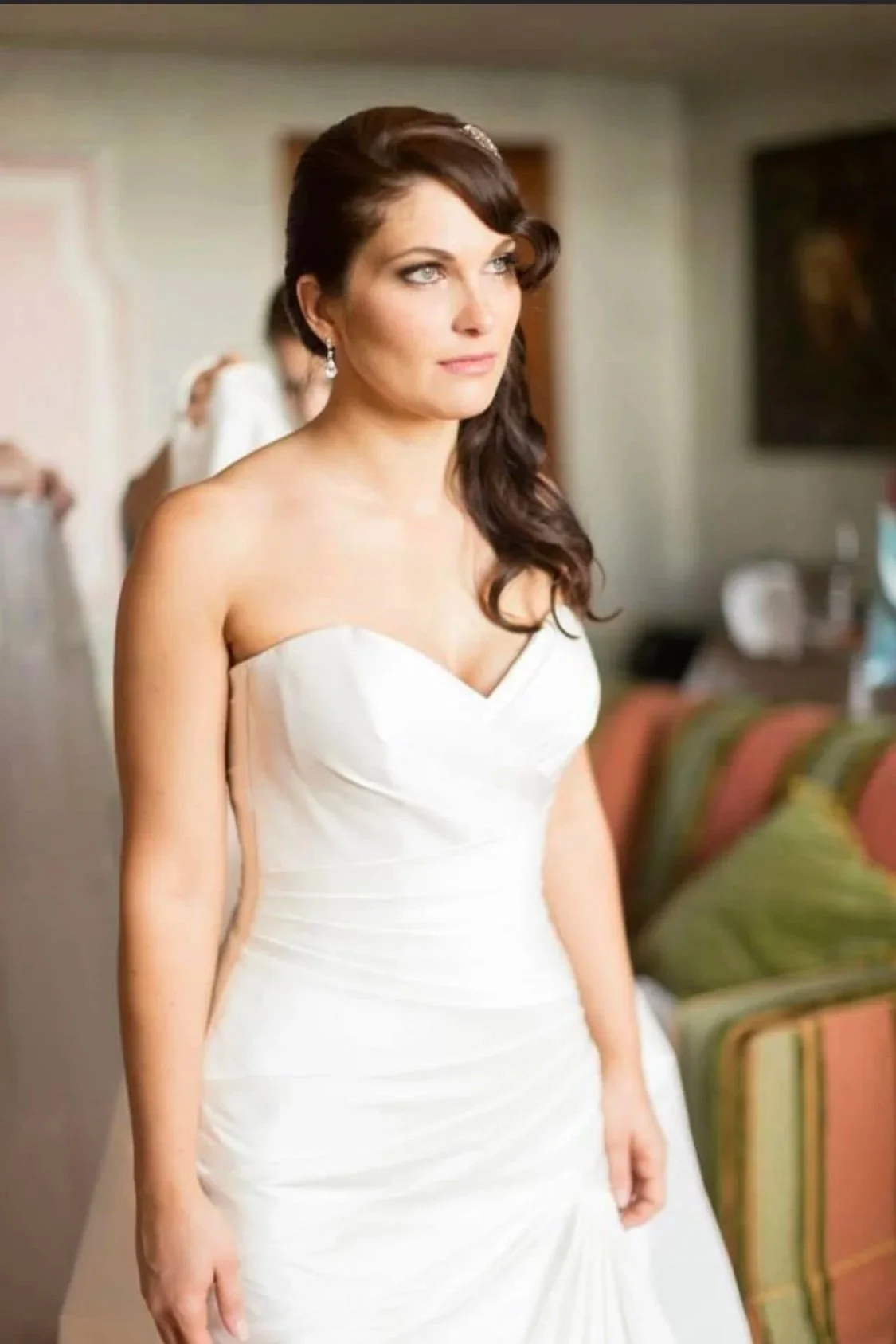A bride with long dark hair styled in loose curls, and classic makeup by the one and only Cecilia Acquaroli.. is wearing a strapless white wedding gown with a sweetheart neckline, standing in a room with blurred background of sofas and other people.