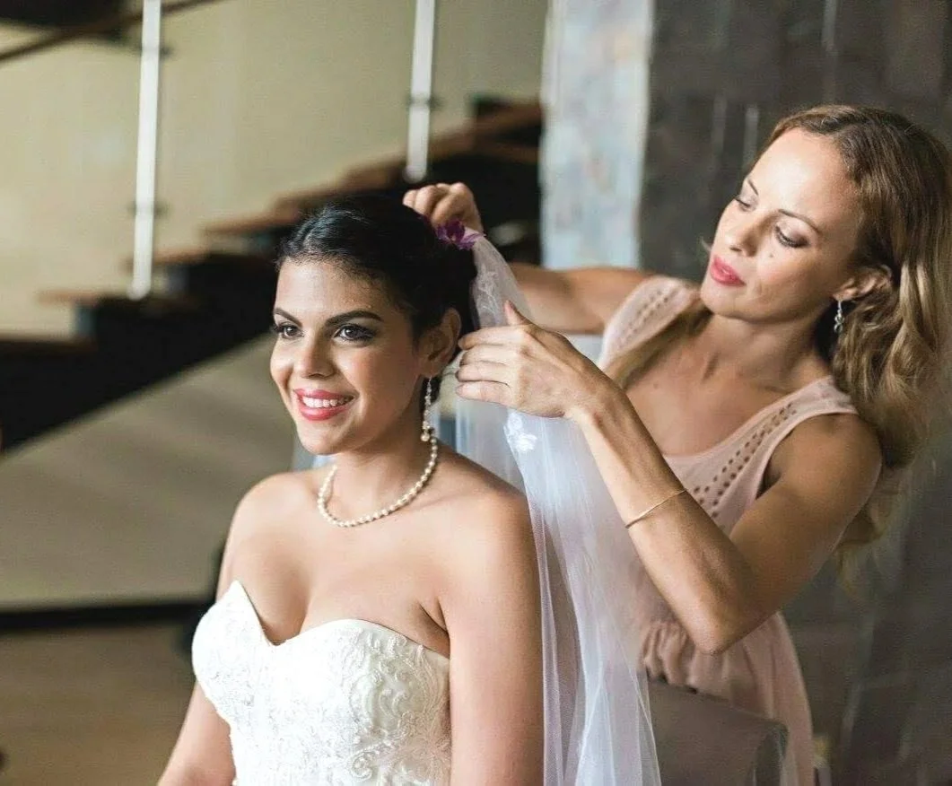 A bride in a strapless wedding dress, smiling as her bridal stylist Cecilia Acquaroli adjusts her veil.