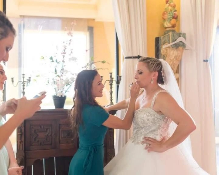 Bride getting her makeup touched up by a bridal stylist Cecilia Acquaroli, while two other women look on, inside a well-lit room with curtains and a wooden sideboard, in Rome, Italy