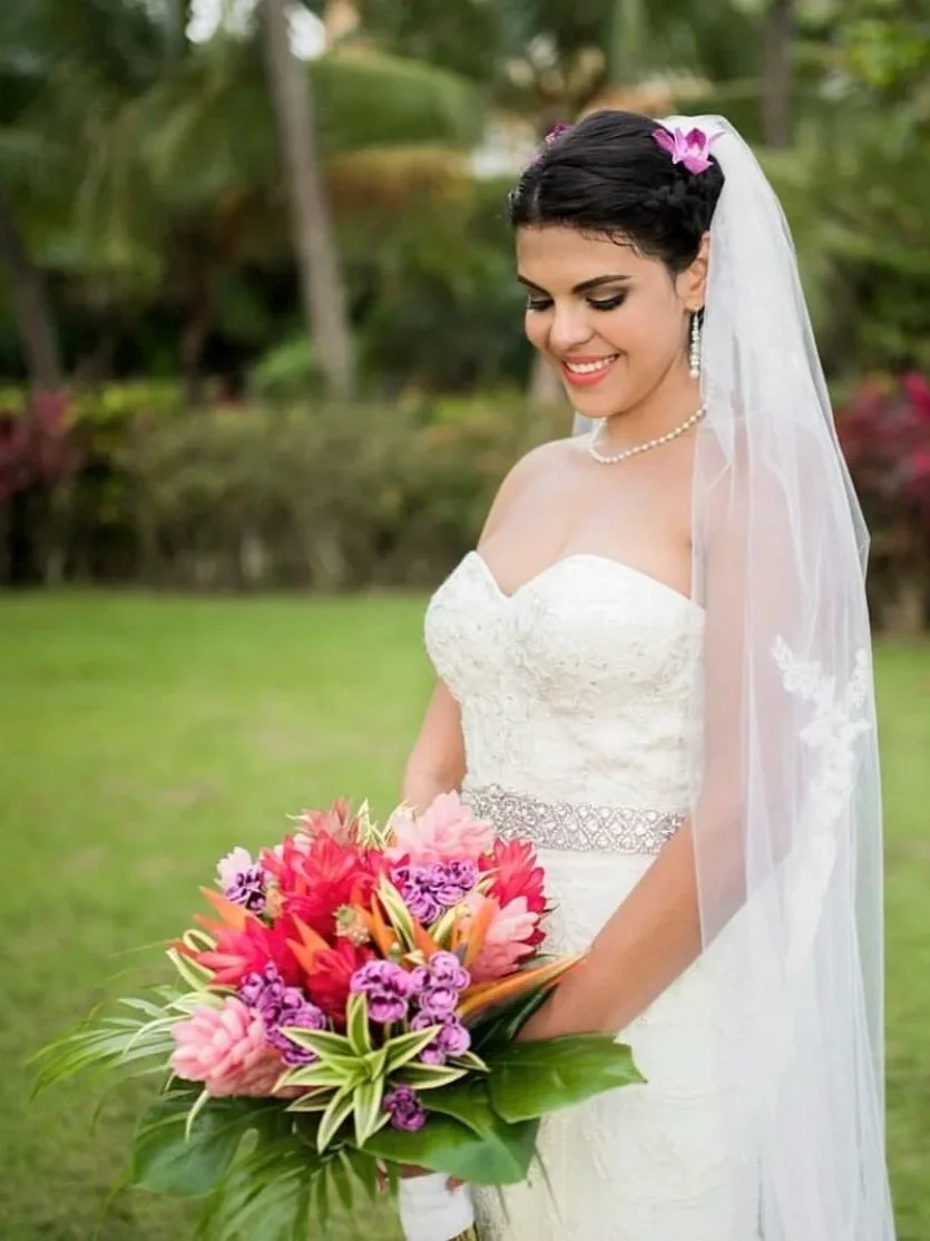 A happy bride in a white wedding dress holding a colorful bouquet of pink, purple, and orange flowers, standing outdoors with greenery and trees in the background, smiling. Hair and makeup by the exceptional Cecilia Acquaroli