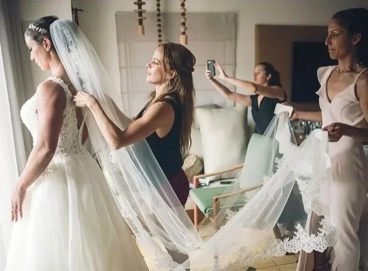 A bride preparing for her wedding, surrounded by women helping with her veil and dress, in a well-lit room.