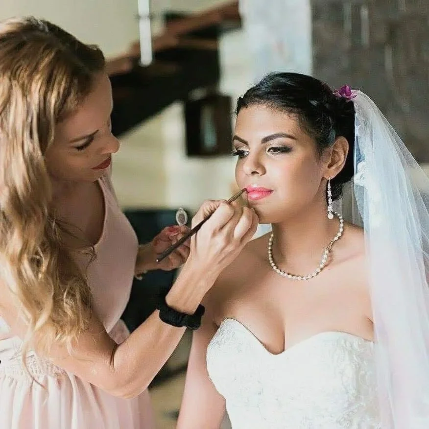 A bride with dark hair styled in an updo, wearing a white strapless wedding gown, pearl necklace, and veil, receives makeup application from  makeup artist  and bridal stylist Ceciia Acquaroli, with long wavy hair, in an indoor setting.