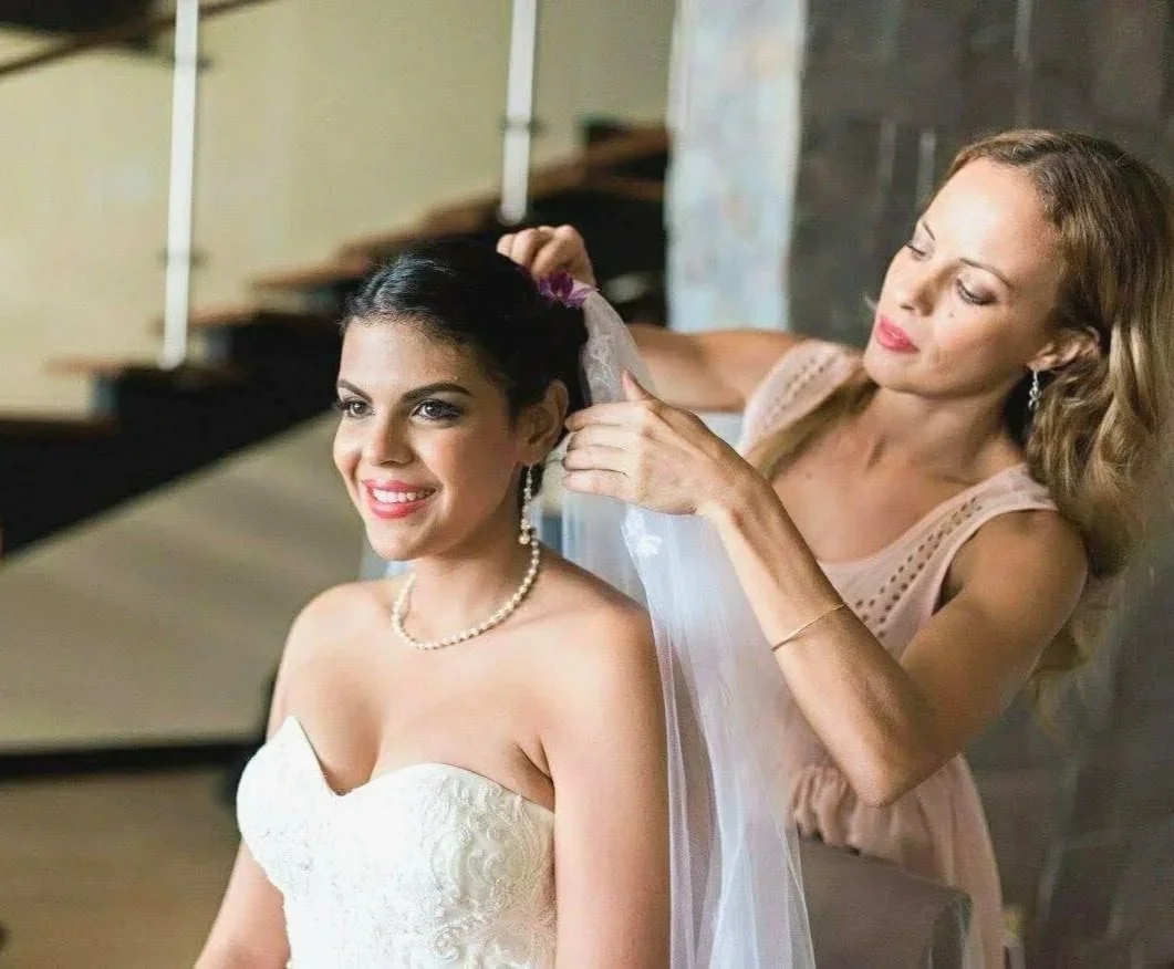 A bride in a strapless wedding dress, smiling as her bridal stylist Cecilia Acquaroli adjusts her veil.