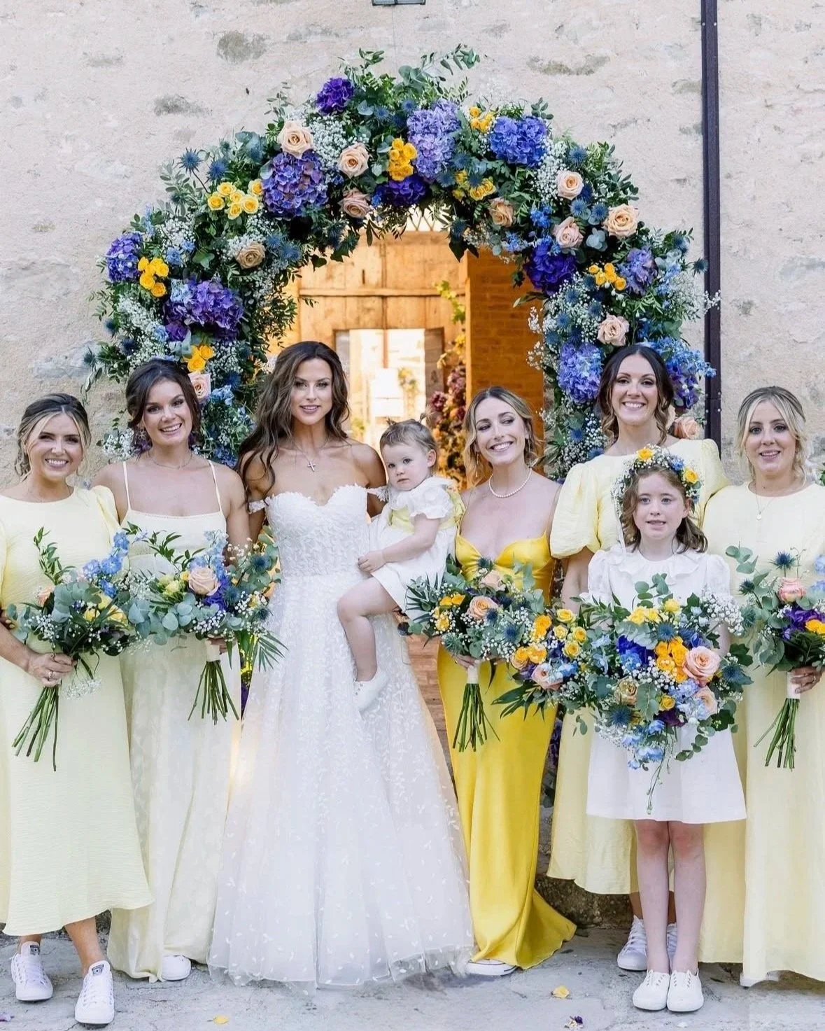 A group of women and children celebrating at a wedding or special event, standing under a large floral archway decorated with purple, blue, yellow, and white flowers. The women are dressed in yellow and white outfits, holding bouquets, and some have 