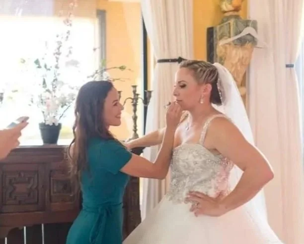 Bride getting her makeup touched up by a bridal stylist Cecilia Acquaroli, while two other women look on, inside a well-lit room with curtains and a wooden sideboard, in Rome, Italy
