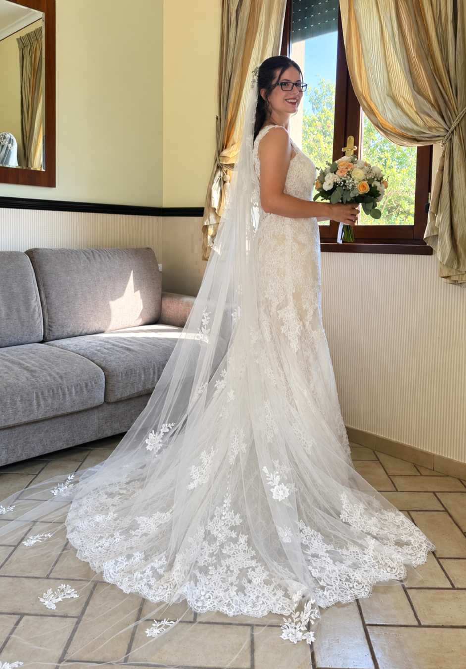 Bride in a lace wedding dress holding a bouquet, standing by a window with golden curtains, indoors