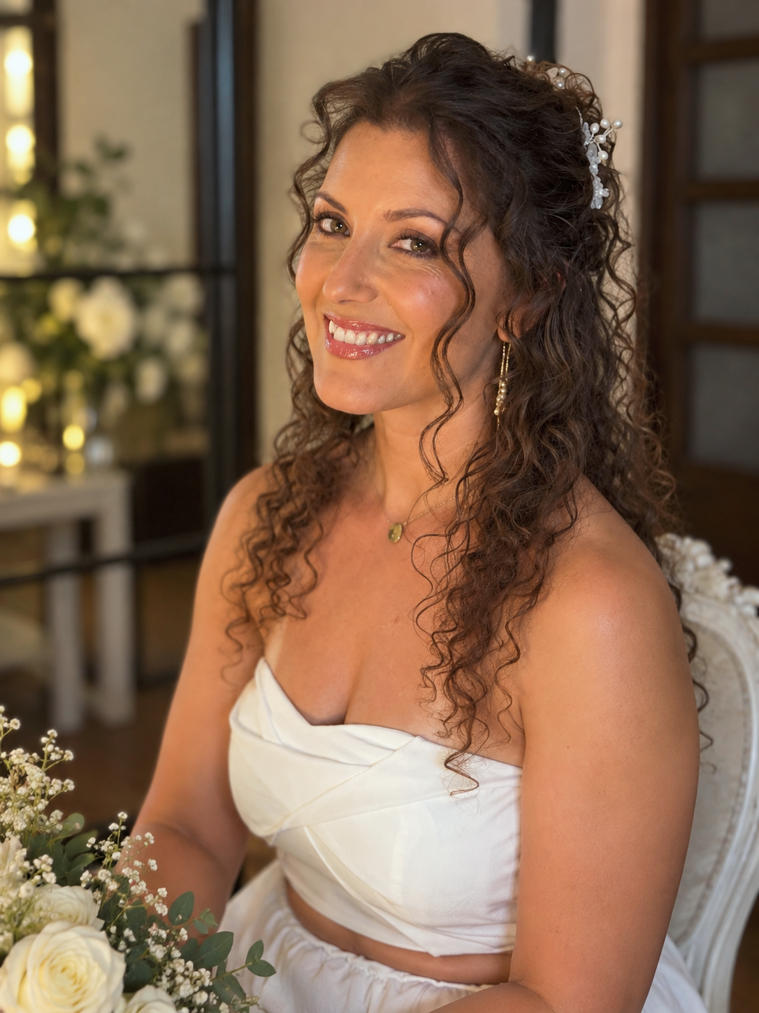 A woman with long, curly brown hair wearing a white strapless dress, smiling, with floral accessories in her hair, sitting indoors with floral arrangements in the background.