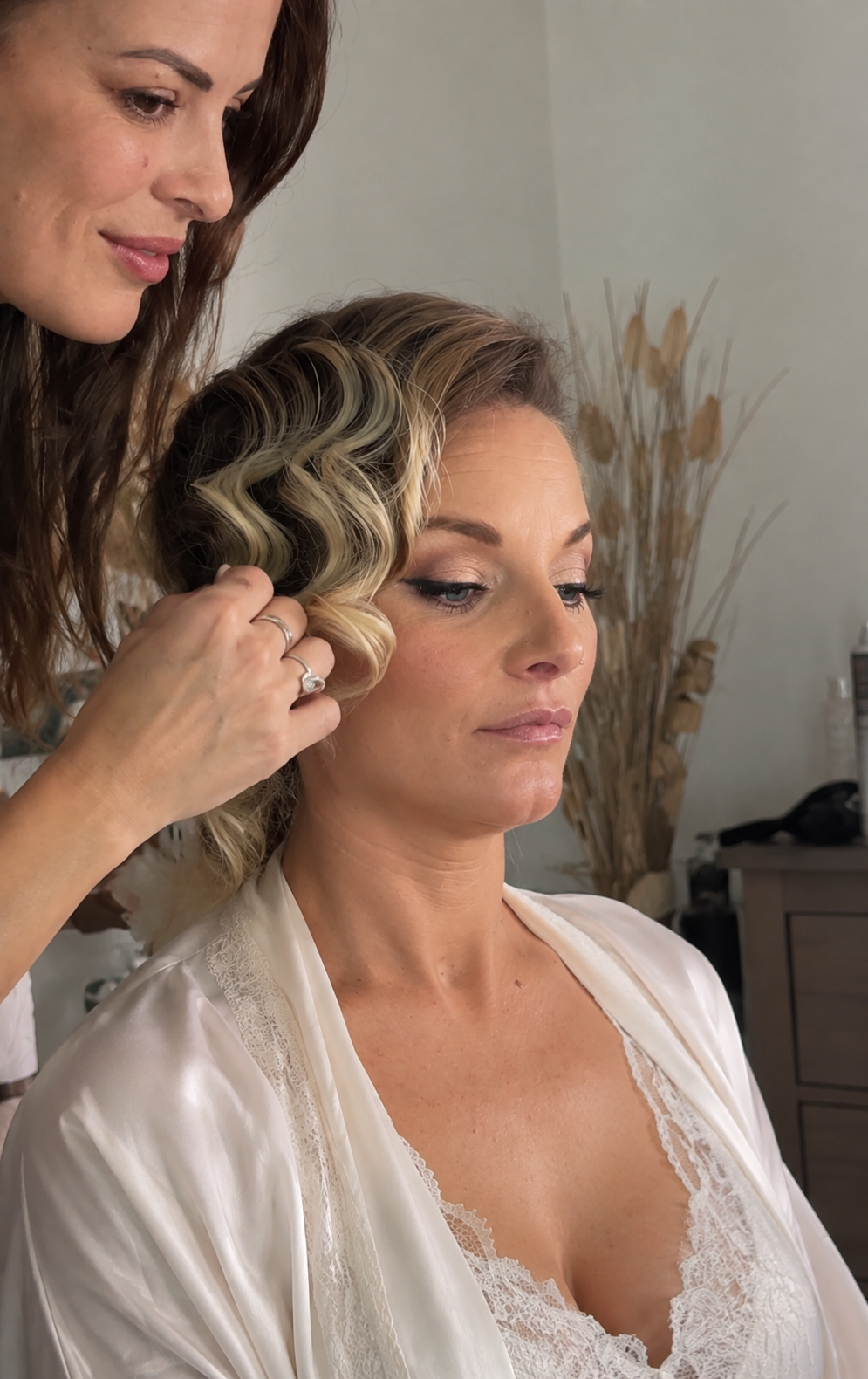 A woman with styled blonde hair wearing a white satin robe sits as a stylist does her hair with large curls, in a room with neutral decor and dried plants in the background.