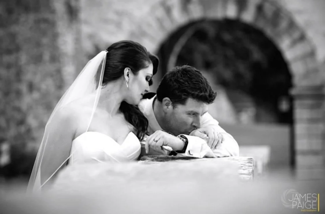 Black and white photo of a bride and groom sitting at a wooden surface, with the bride leaning close to the groom, who is resting his head and arms on the surface, looking contemplative.