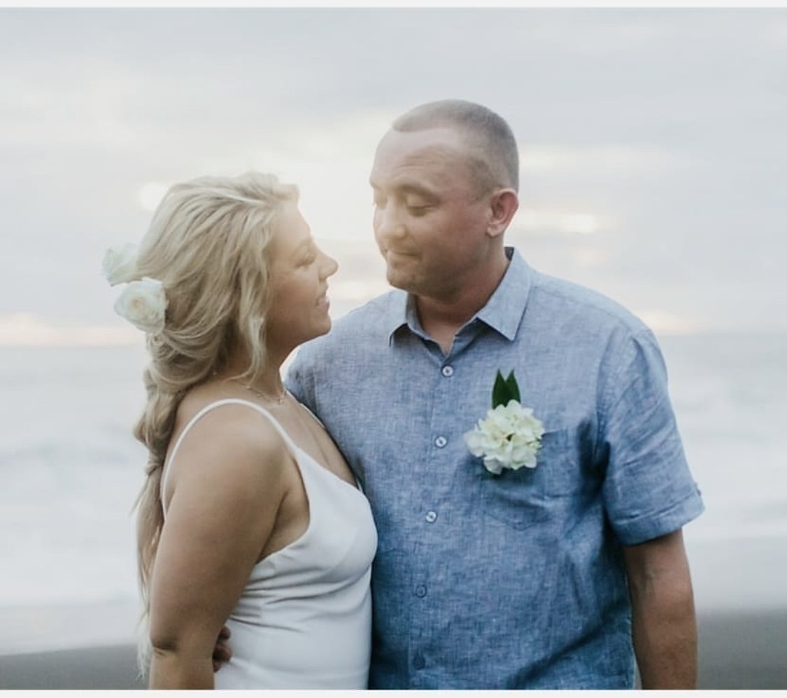 A couple standing close together outdoors near the beach, with the woman wearing a white dress and the man wearing a blue button-up shirt with a white flower boutonniere.