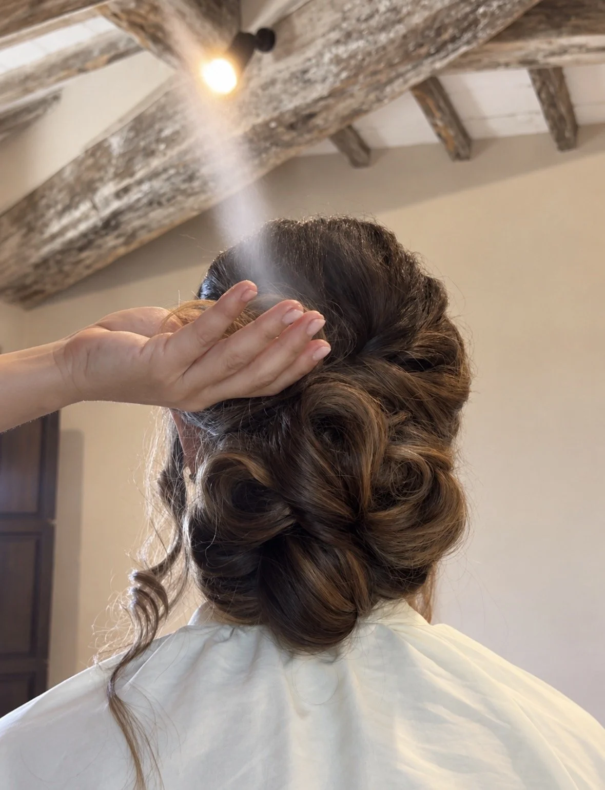 A person with styled, curled hair, seen from behind, getting their hair brushed or styled by someone else in a room with a rustic wooden ceiling.