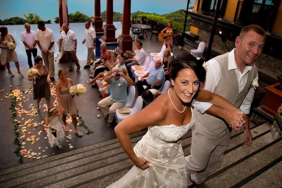 A bride and groom are smiling and holding hands while ascending stairs at their wedding reception. Guests are seated and standing in the background, with some taking photos. Petals are scattered on the floor, and a couple of flower girls are visible.