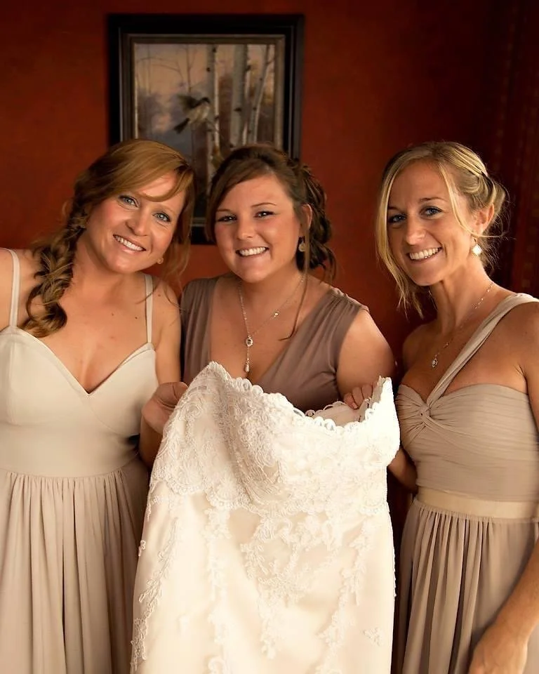 Three women in beige dresses smiling, holding a lace wedding dress, standing in a warmly lit room with a framed picture of trees on the wall behind them.