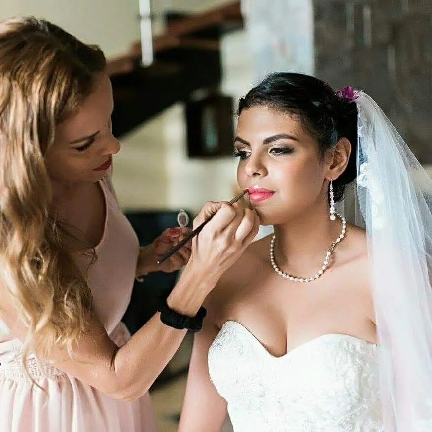A bride with dark hair styled in an updo, wearing a white strapless wedding gown, pearl necklace, and veil, receives makeup application from  makeup artist  and bridal stylist Ceciia Acquaroli, with long wavy hair, in an indoor setting.