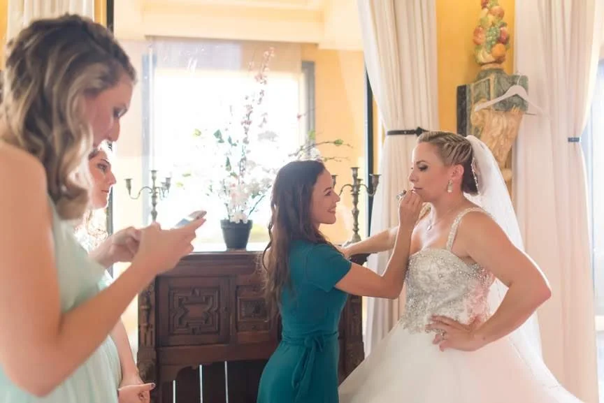 Bride getting her makeup touched up by a bridal stylist Cecilia Acquaroli, while two other women look on, inside a well-lit room with curtains and a wooden sideboard, in Rome, Italy