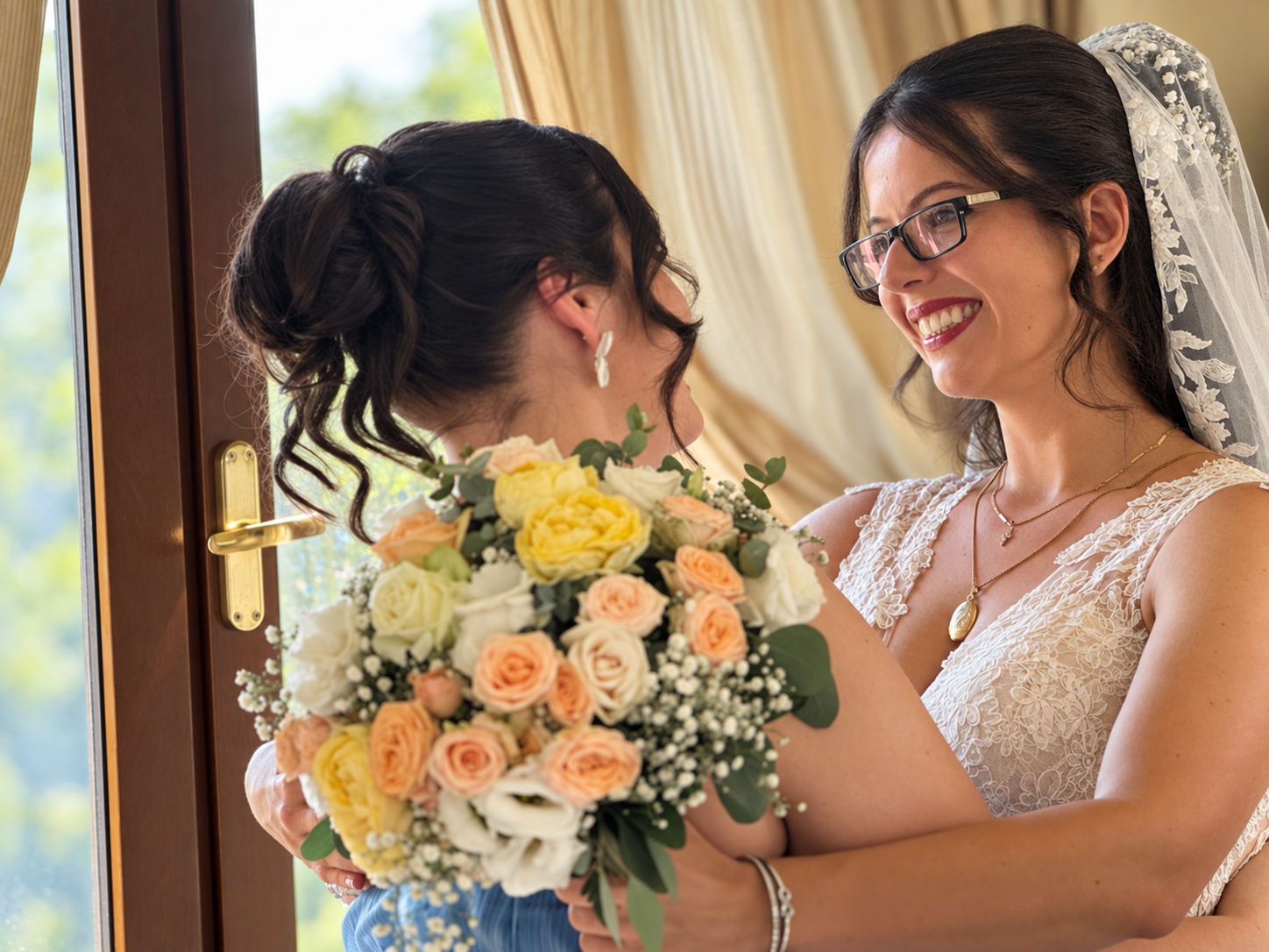 Two women, one in a wedding dress wearing a veil and glasses, and the other with curly dark hair and earrings, share a moment indoors during a wedding, with a bouquet of flowers.