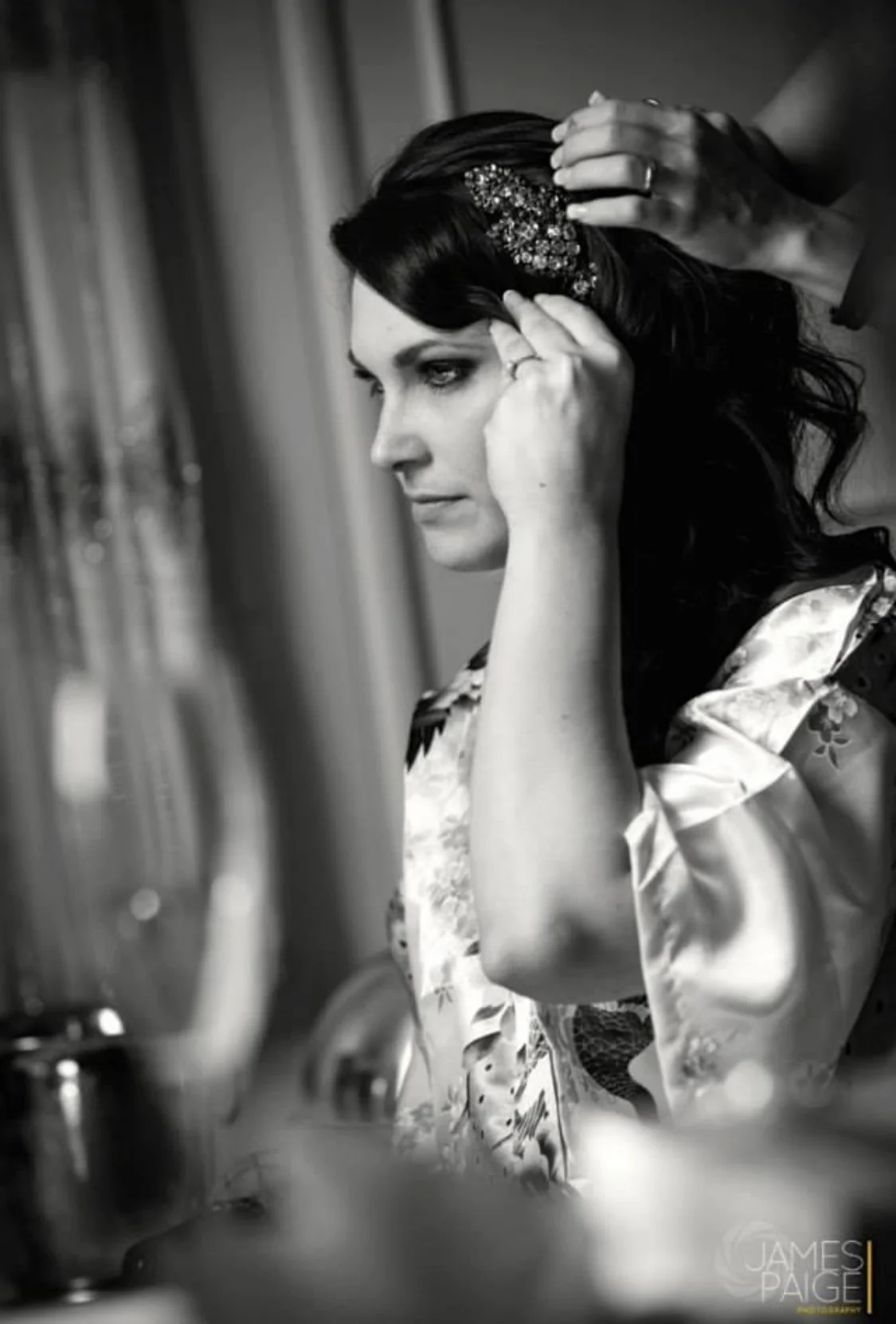 A black and white photo of a woman with dark hair getting ready, possibly for a special occasion. Someone is fixing her hair, which is adorned with a decorative hairpiece. She is dressed in silk or satin robes and appears to be sitting in front of a 