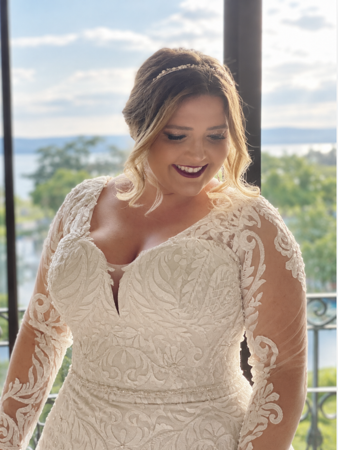 A smiling woman in a cream lace wedding dress with floral embroidery, standing inside near a window overlooking water and trees.
