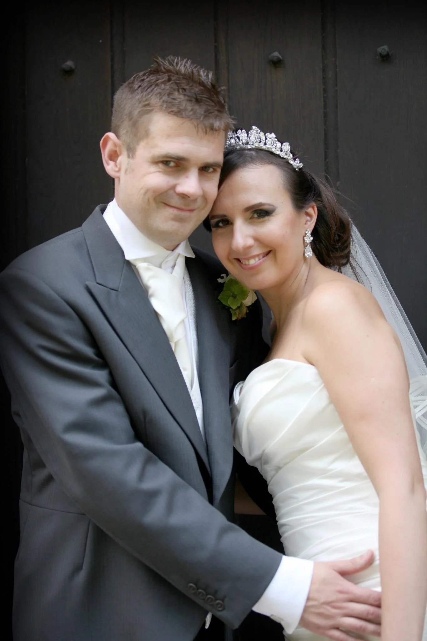 A newlywed couple smiling, the groom in a gray suit and white tie, and the bride in a strapless white wedding dress with a jeweled tiara and earrings, standing in front of a dark wooden wall.