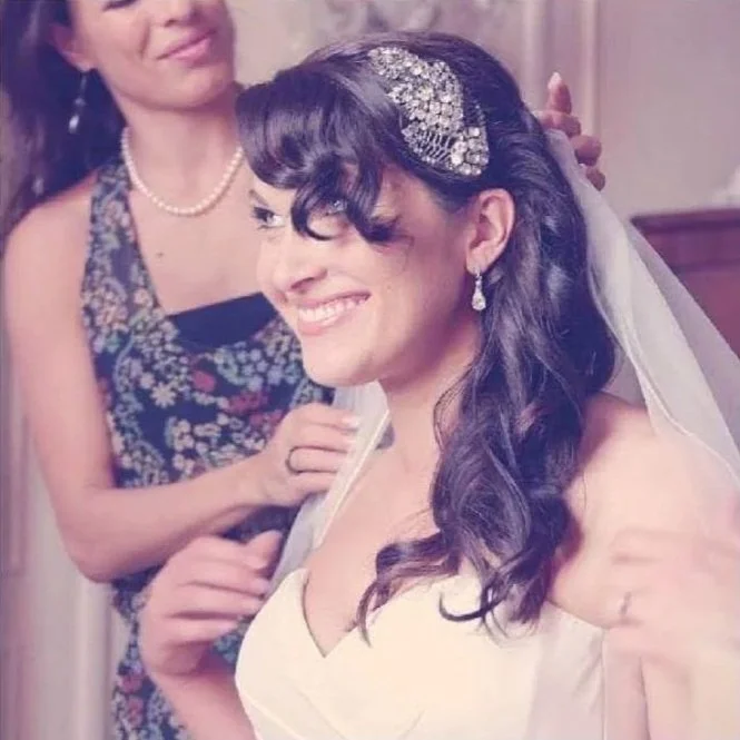 A bride with dark, curled hair and a sparkling hairpiece, smiling, with Cecilia Acquaroli behind her helping with her wedding attire and final details before the big day