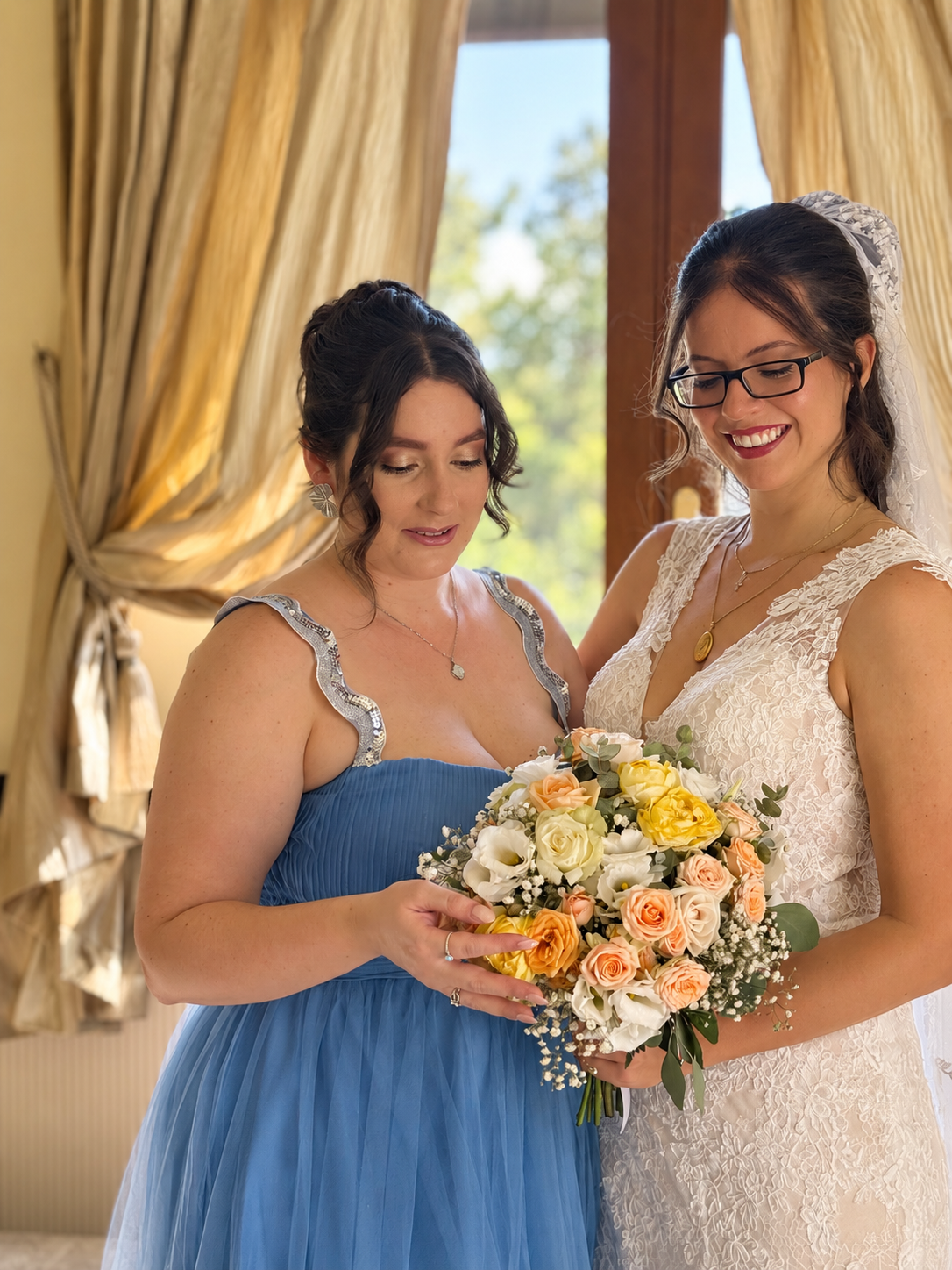 A bride and woman in a blue dress, possibly a bridesmaid, share a moment holding a bouquet of flowers inside a sunny room with yellow curtains and a large window.