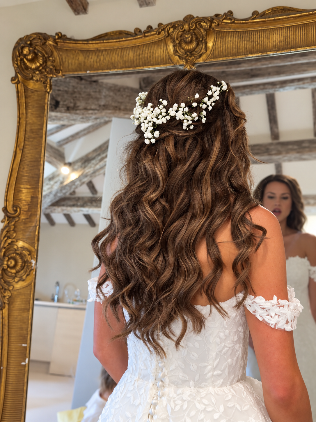 A woman with long, curly brown hair wearing a floral headband and a white lace wedding dress looking into a mirror.