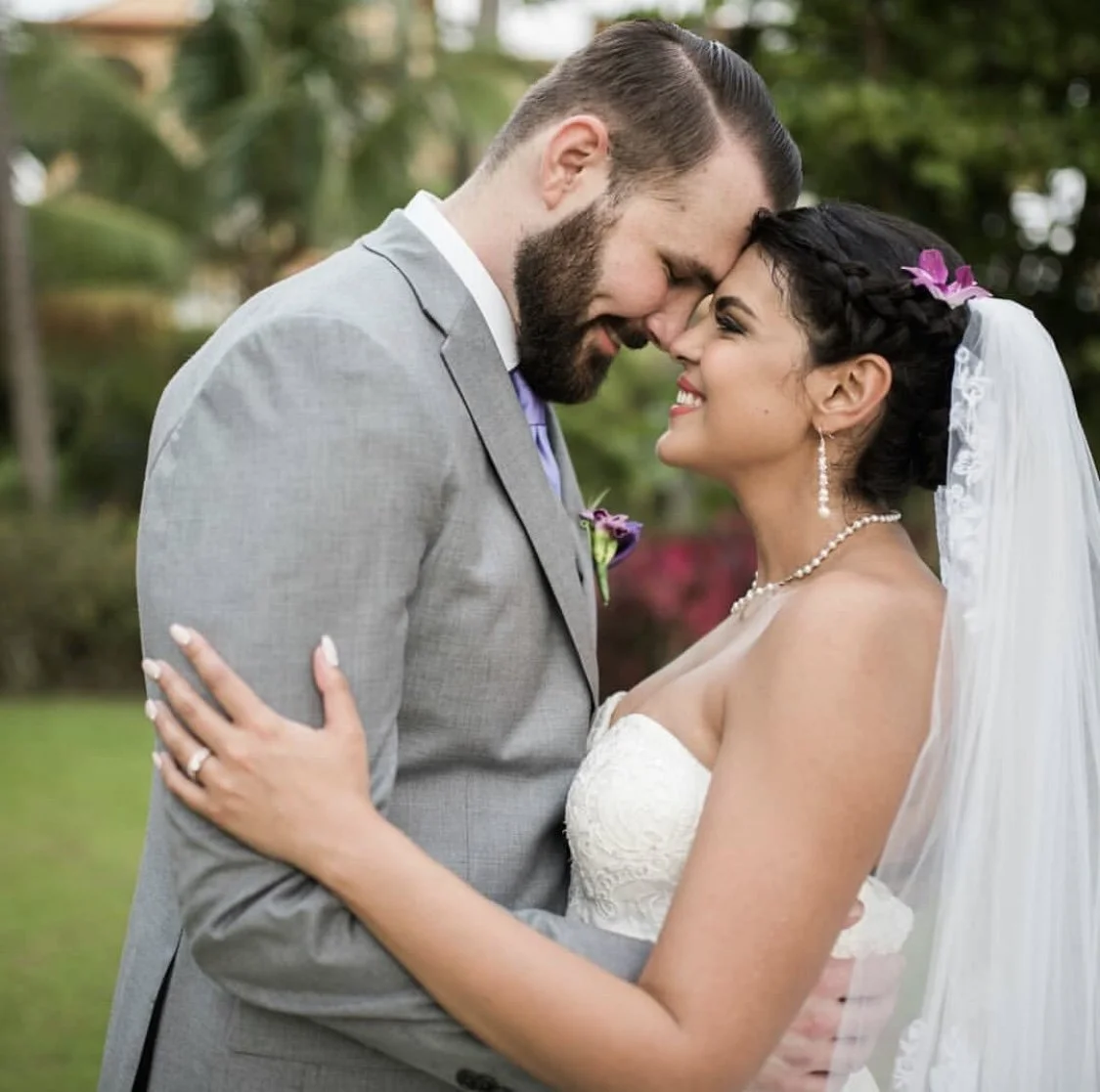 A bride and groom share a tender moment outdoors, touching foreheads and smiling, with lush green foliage in the background.