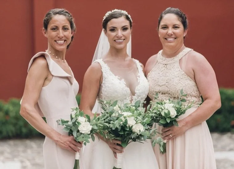 Three women standing outside against a red wall, celebrating a wedding. The bride is in the middle wearing a white wedding dress with lace and a veil, holding a bouquet of white roses and greenery. The women on each side of her are dressed in light-c