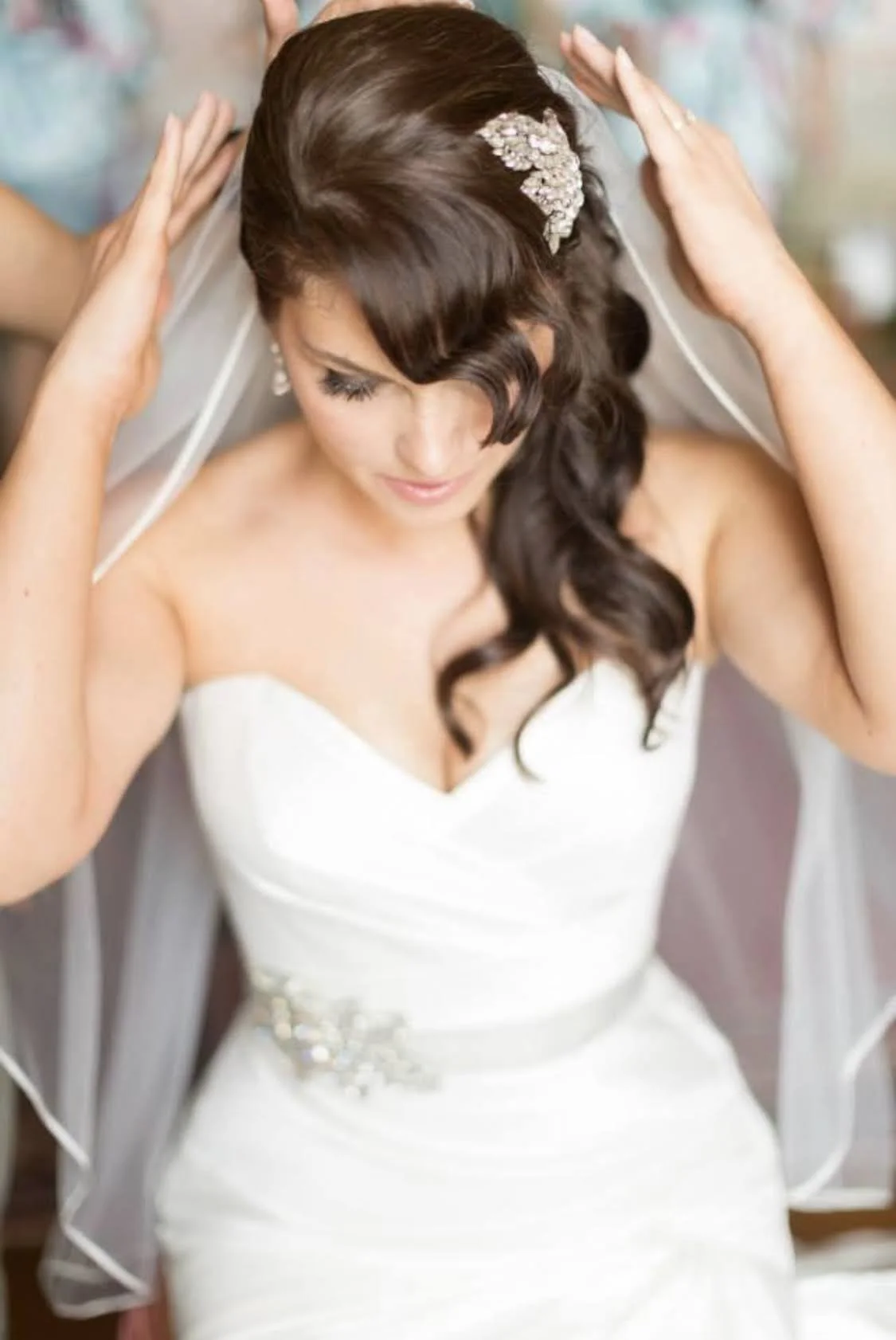 A bride with dark brown, wavy hair and a jeweled hair accessory, adjusting her veil before a wedding. Styled by Cecilia Acquaroli