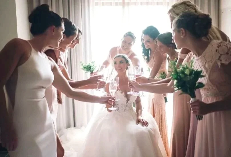A bride in a wedding dress surrounded by her bridesmaids holding champagne glasses in a celebratory toast.