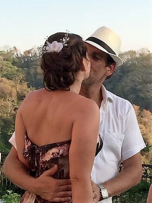 A couple sharing a kiss outdoors, with the woman wearing a strapless dress and the man wearing a white shirt and a white fedora hat. featuring soft and natural wedding styling by professional artist Cecilia Acquaroli.