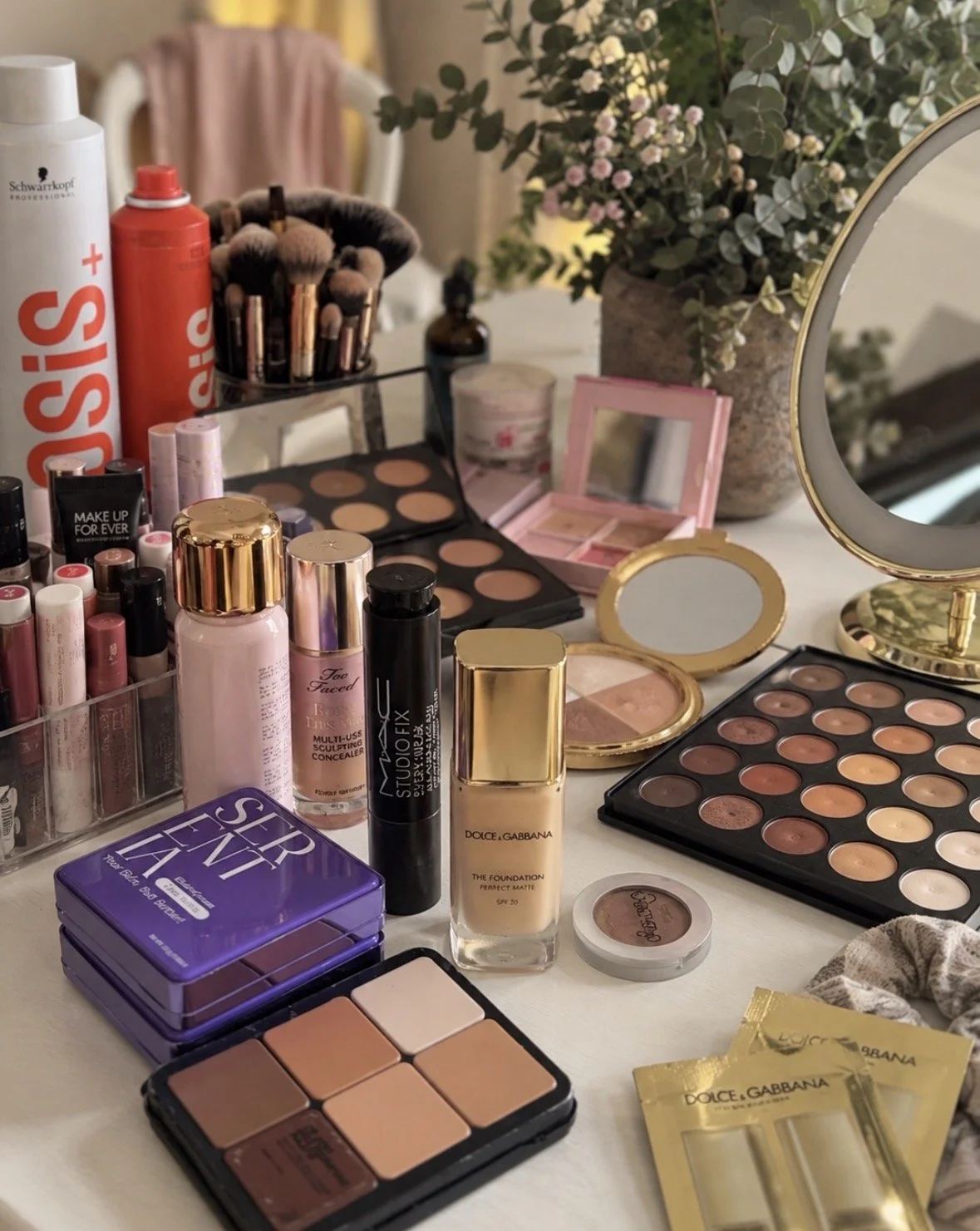 A makeup vanity table with various beauty products including foundation, lipsticks, eyeshadow palettes, brushes, and skincare items, along with a large flower arrangement in the background.