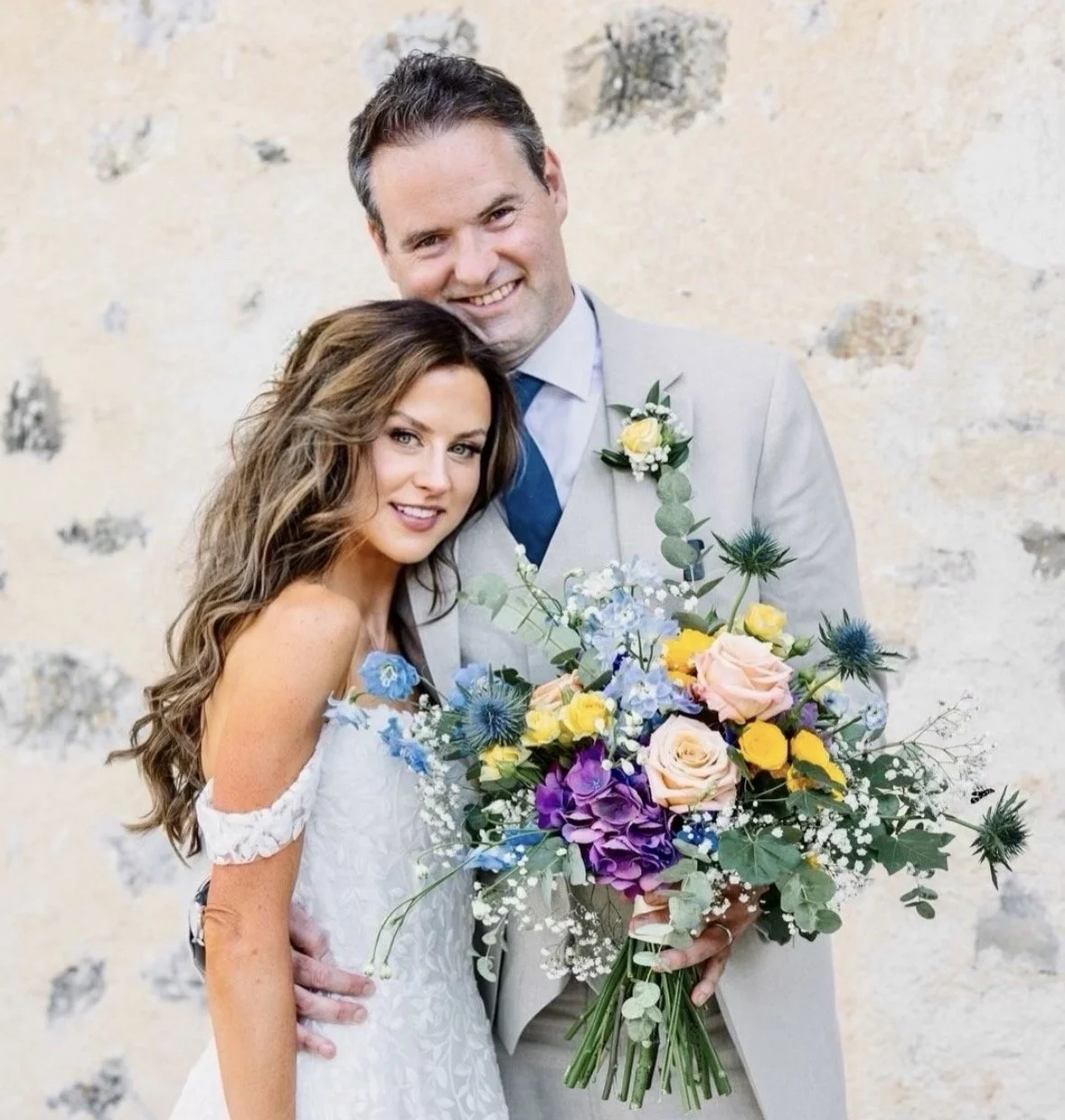 A bride and groom smiling, holding a colorful bouquet of flowers, standing in front of a textured beige wall.