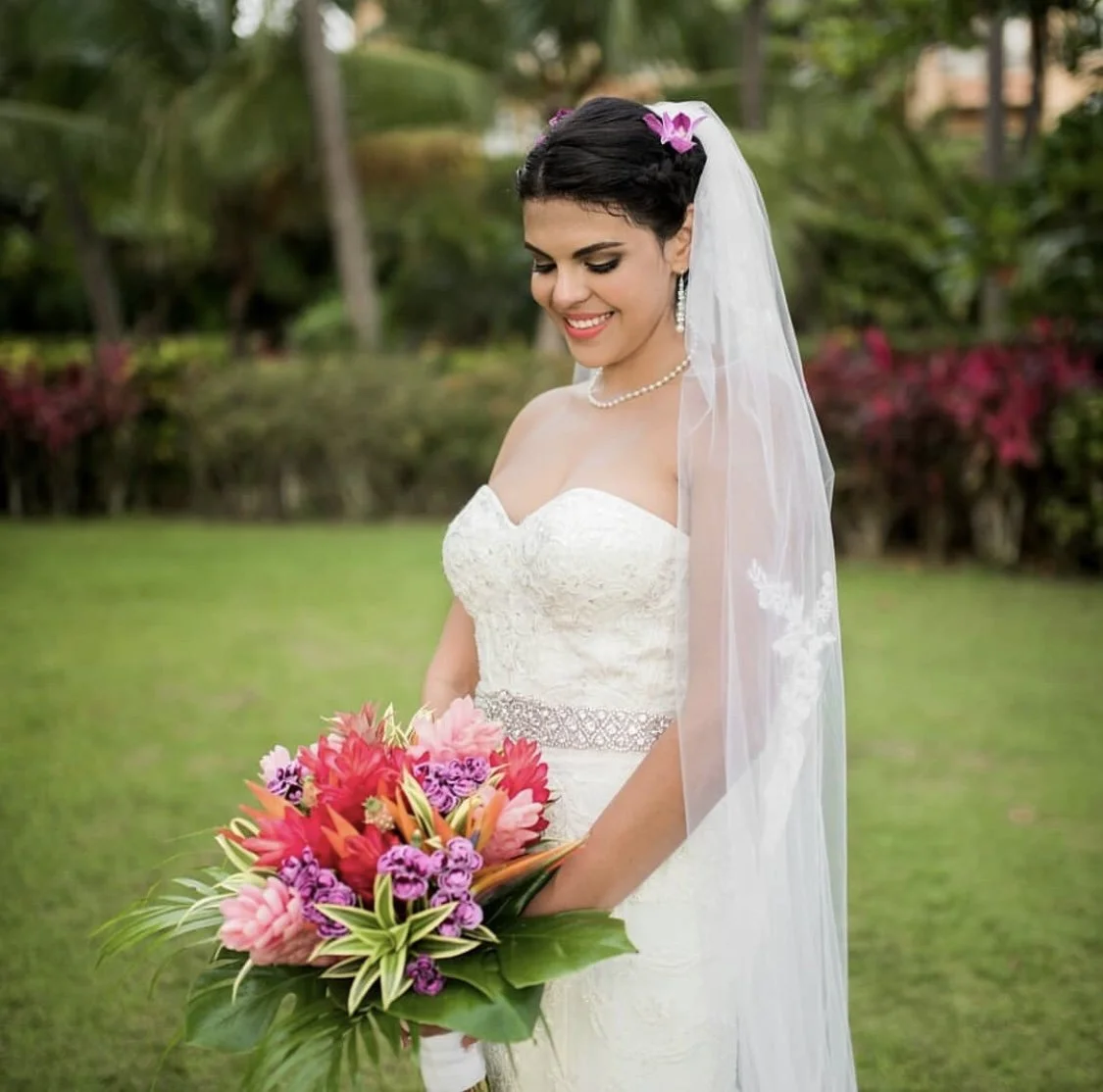 A happy bride in a white wedding dress holding a colorful bouquet of pink, purple, and orange flowers, standing outdoors with greenery and trees in the background, smiling. Hair and makeup by the exceptional Cecilia Acquaroli