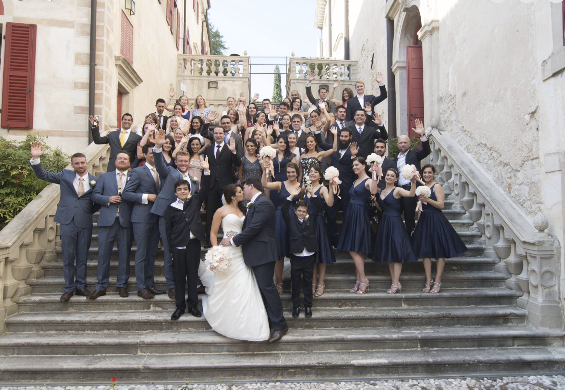 A large wedding party on a staircase outside a historic building. The bride and groom are in the center, holding hands, with the bride in a white wedding gown holding a bouquet of flowers. The bridesmaids are dressed in navy blue dresses, and the groomsmen are in suits. Everyone is waving and smiling for the photo.