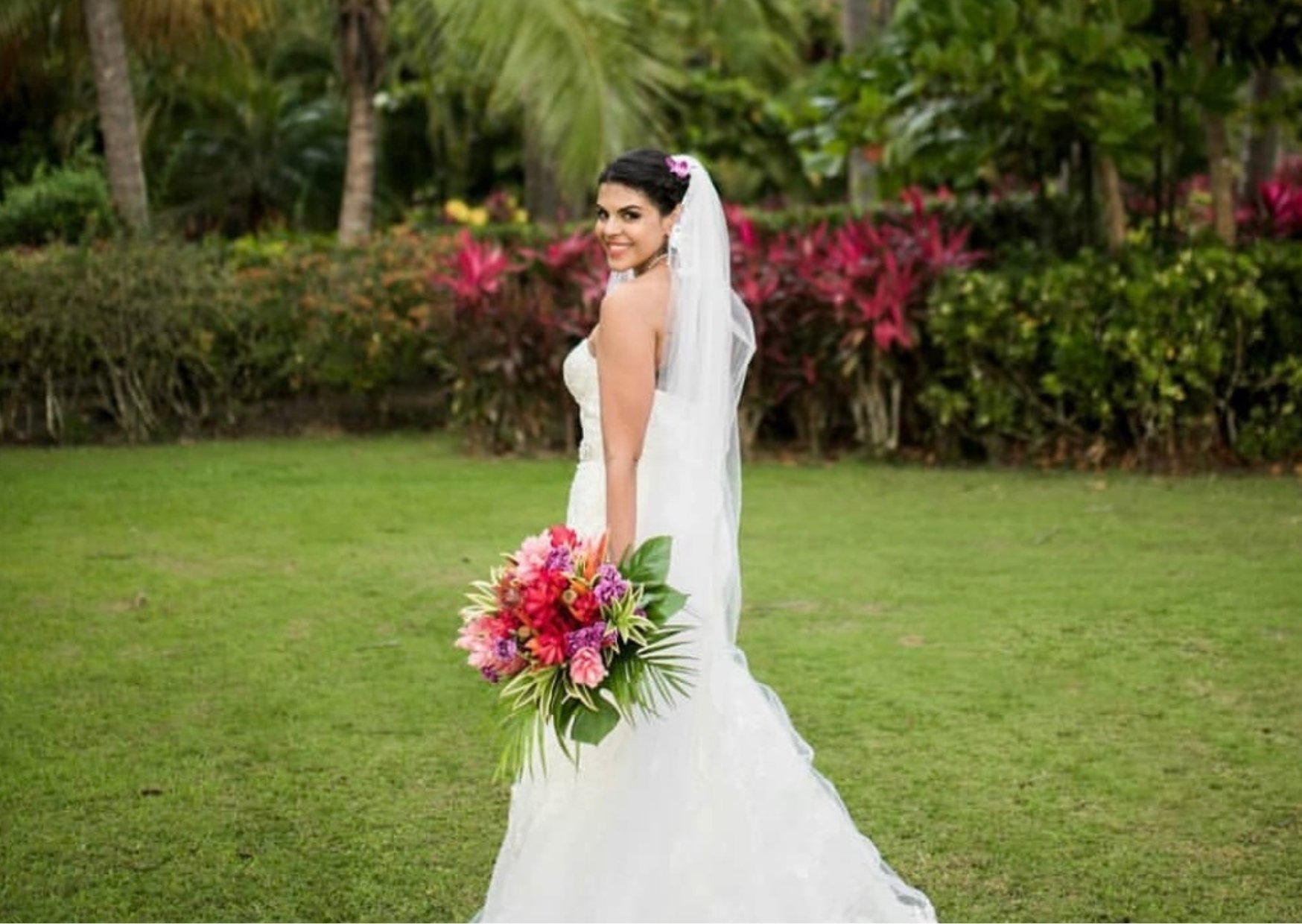 A smiling bride in a white wedding gown holding a vibrant bouquet of pink, red, and purple flowers on a lush green lawn with tropical plants in the background.