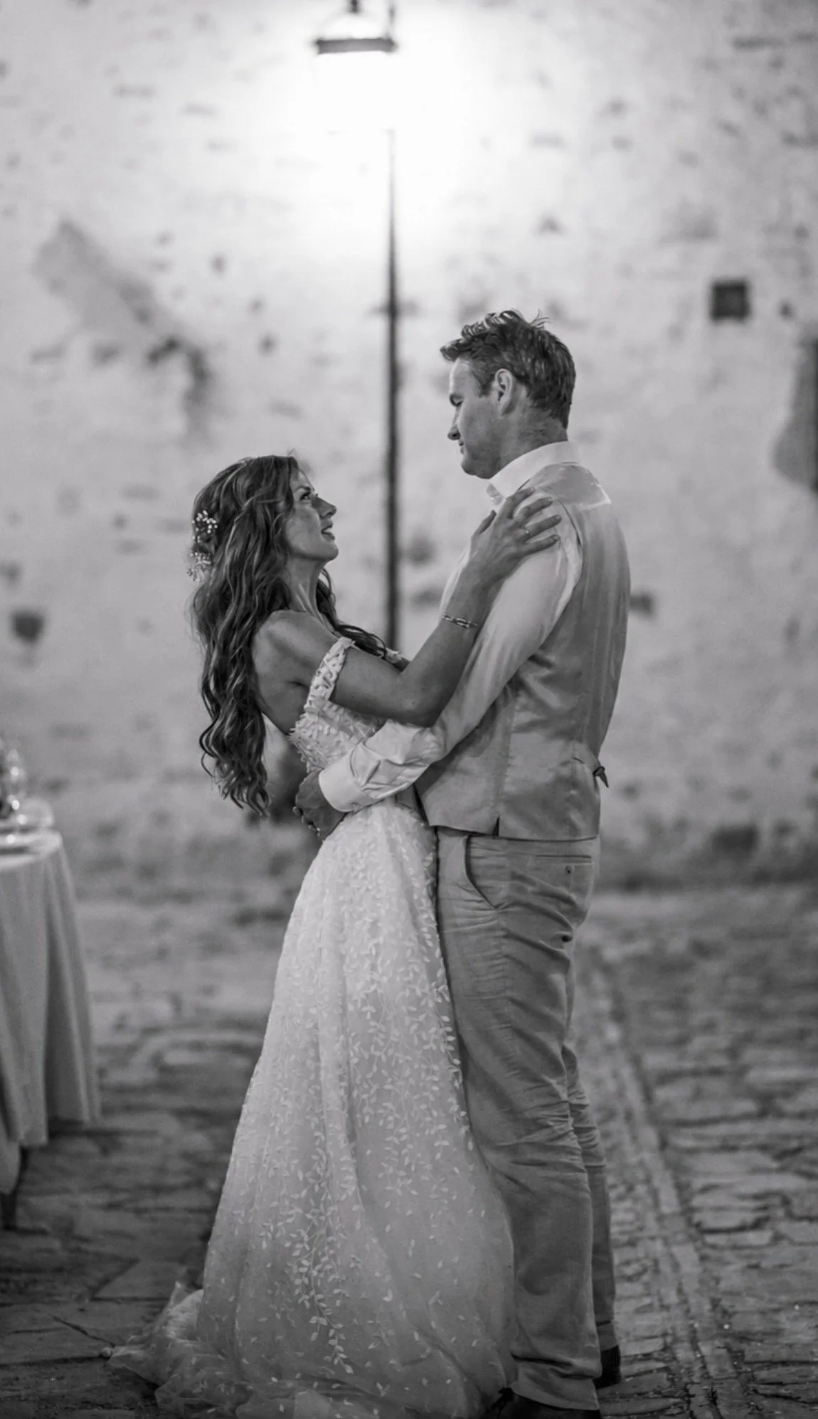 A black-and-white photo of a bride and groom dancing at their wedding reception. The bride is wearing a lace wedding gown and the groom is in a light-colored vest and pants. They are looking at each other with affection, holding close in front of a b