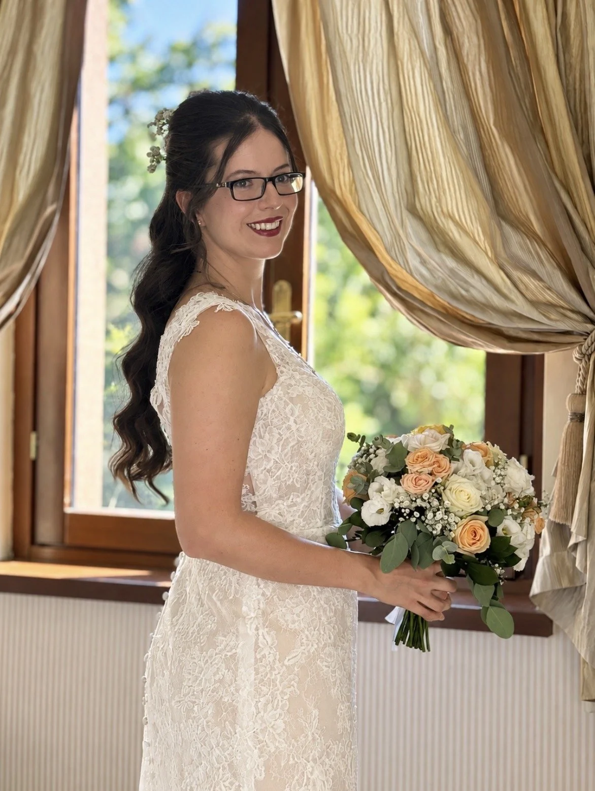 A bride in a lace wedding dress holding a bouquet of peach and white roses, standing indoors near a window with golden curtains.