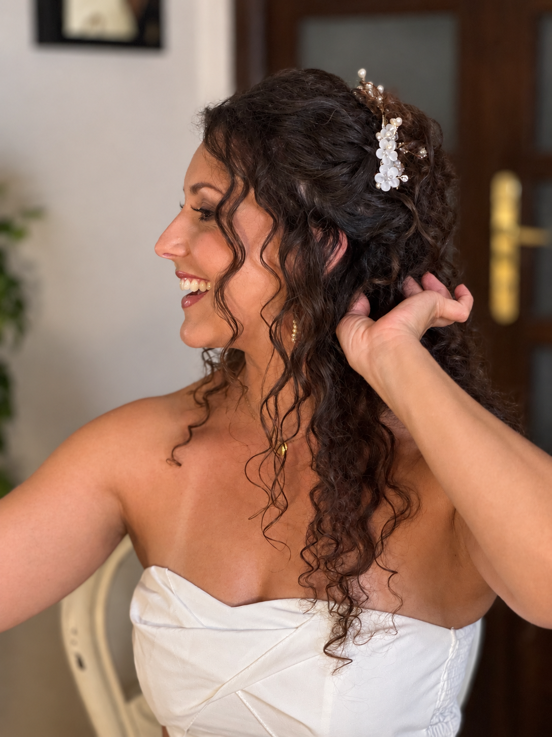 A woman with curly hair, decorated with a floral hairpiece, smiling and touching her hair, wearing a strapless white dress.