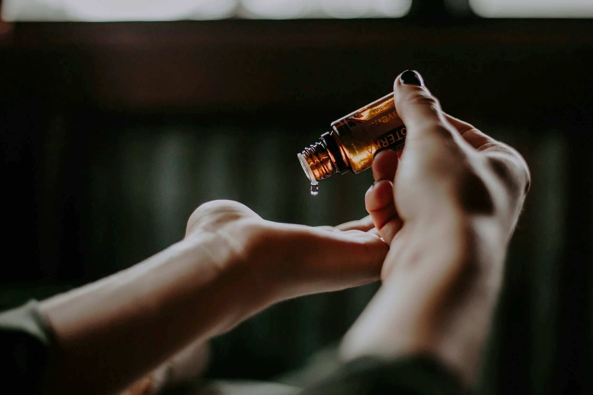 A person's hands holding a small glass bottle of essential oil, with a drop of oil falling from the opening.