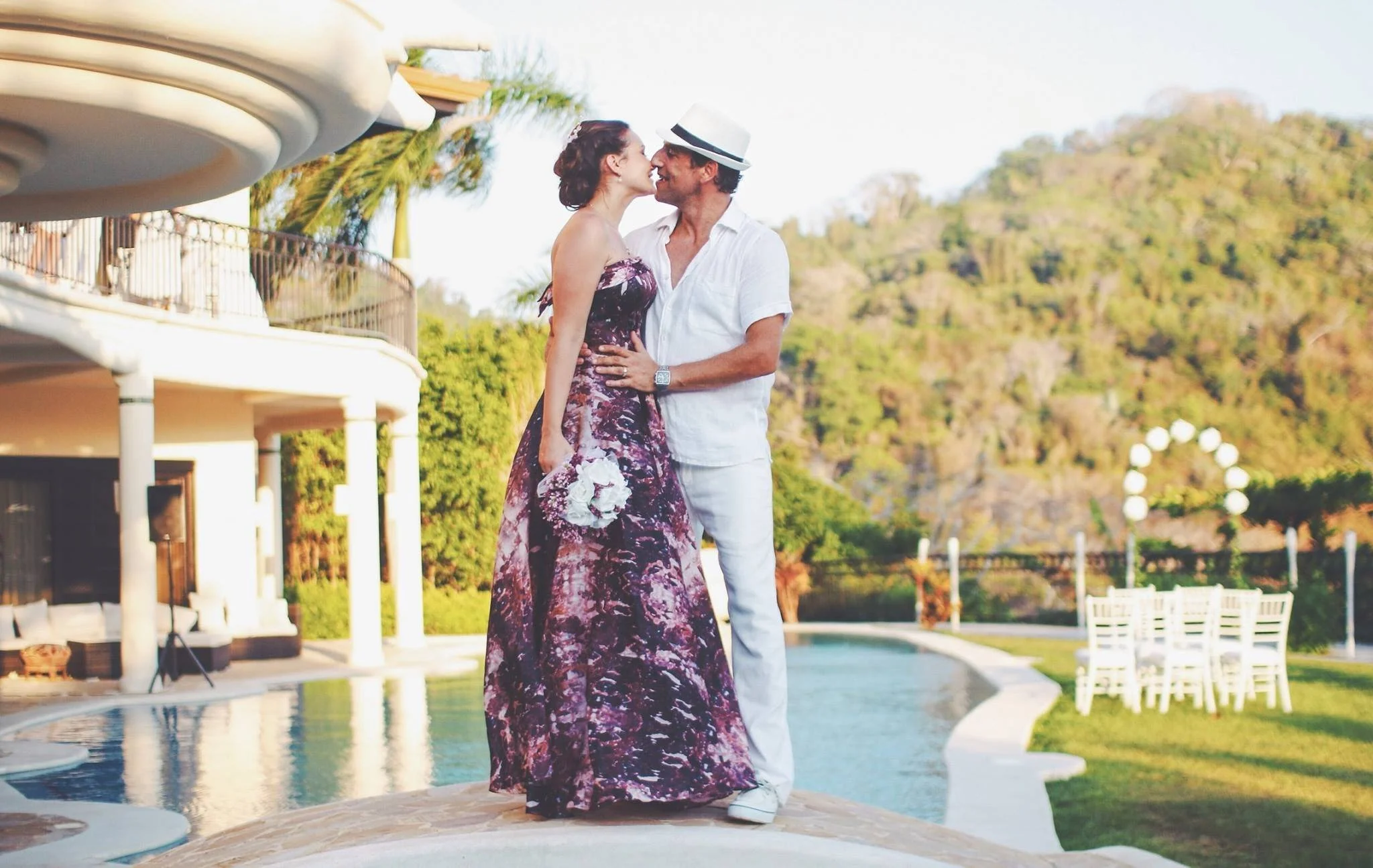 A couple dressed in summer attire, sharing an intimate moment, standing by a swimming pool outdoors with lush greenery and hills in the background, with expert on-location beauty services provided by Cecilia Acquaroli