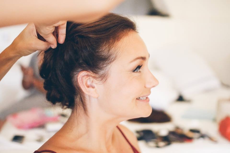A woman getting her hair styled by a person whose hand is visible, with a blurred background of a bed and various personal items.