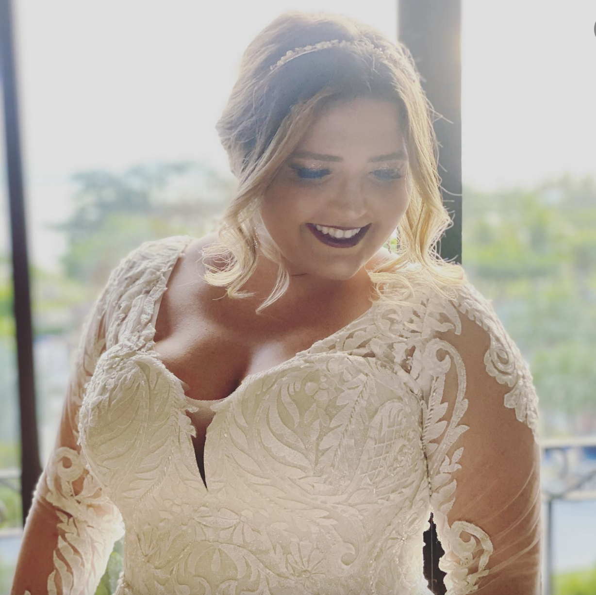 A smiling woman in a cream-colored lace dress with detailed embroidery, standing by a window with greenery outside.