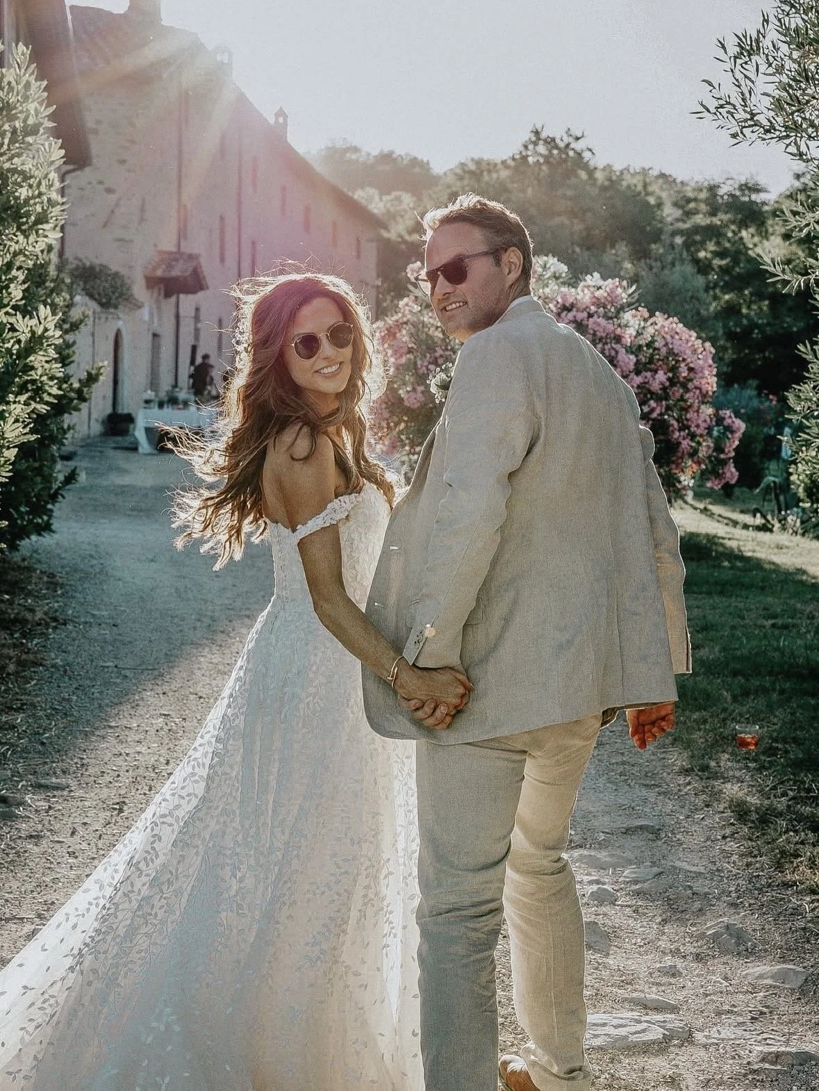 A smiling woman in a white wedding dress and sunglasses holding hands with a man in a light-colored suit holding a small flower, outdoors on a sunny day with greenery and pink flowering bushes in the background.