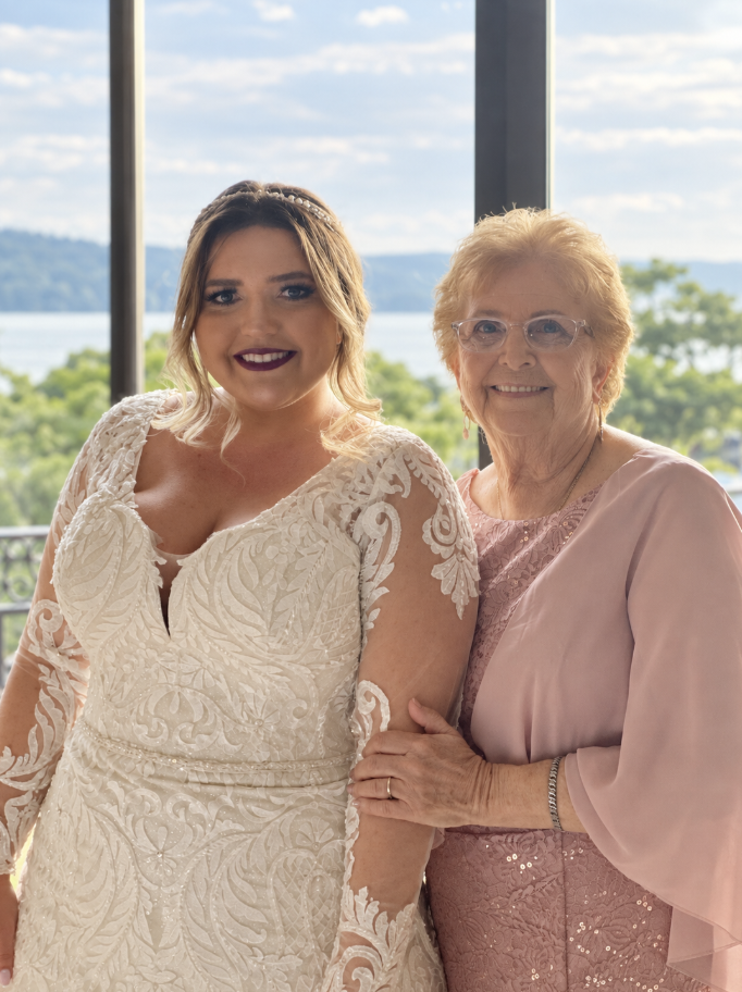 A young woman in a white lace wedding dress stands next to an older woman in a pink dress with lace details. They are smiling and standing in front of a window with a scenic view of water and greenery.
